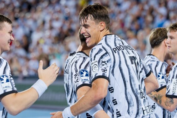 Eine Gruppe männlicher Handballspieler in schwarz-weißen Trikots lächelt und feiert gemeinsam auf dem Spielfeld, während im Hintergrund eine jubelnde Menschenmenge zu sehen ist.