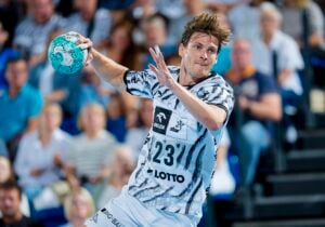Ein männlicher Handballspieler in einem schwarz-weißen Trikot macht sich während eines Spiels bereit, den Ball zu werfen, während im Hintergrund eine verschwommene Zuschauermenge zusieht.