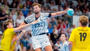 Ein Handballspieler in einem weißen Trikot springt hoch, um einen Wurf zu versuchen, während er von zwei Spielern in gelben Trikots in einem spannenden Spiel verteidigt wird; im Hintergrund beobachtet das Publikum das Geschehen.