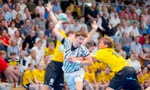 Ein männlicher Handballspieler in einem weißen Trikot versucht während eines spannenden Spiels, an zwei Verteidigern in gelben Trikots vorbeizukommen, während im Hintergrund die Zuschauer zuschauen.