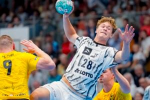Ein männlicher Handballspieler in einem schwarz-weißen Trikot springt hoch, um den Ball zu werfen, während zwei Verteidiger in gelben Trikots versuchen, ihn während eines Spiels zu blockieren; im Hintergrund ist eine verschwommene Zuschauermenge zu sehen.