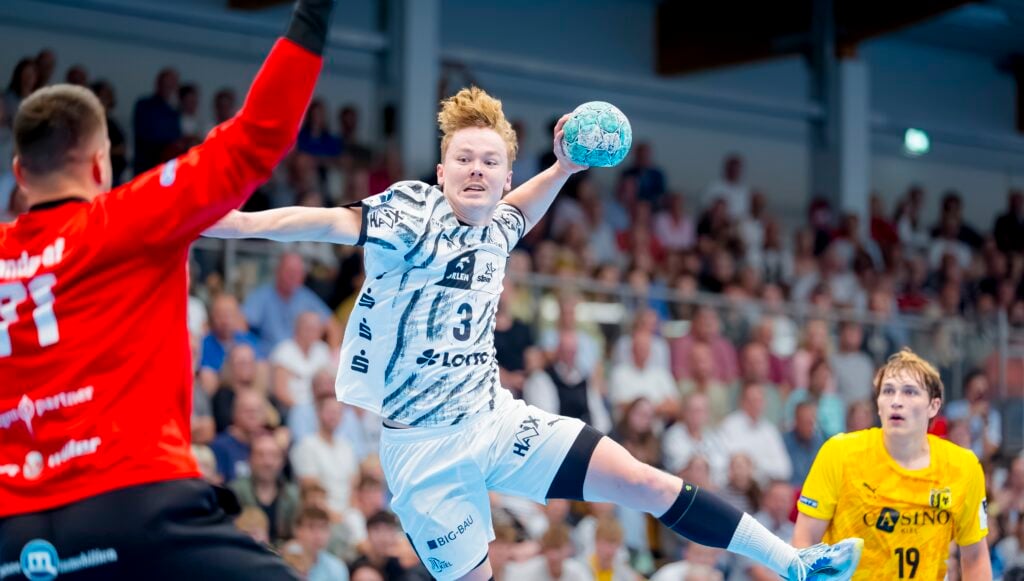 Ein Handballspieler im weißen Trikot springt zum Wurf hoch, während sich ein Torwart in Rot darauf vorbereitet, den Ball abzuwehren. Ein weiterer Spieler in Gelb beobachtet das Geschehen von hinten. Im Hintergrund ist eine Zuschauermenge zu sehen.