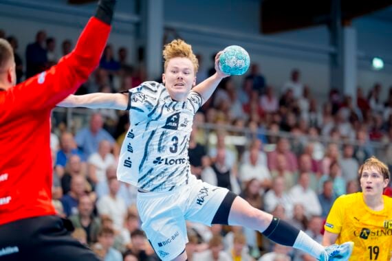 Ein Handballspieler im weißen Trikot springt zum Wurf hoch, während sich ein Torwart in Rot darauf vorbereitet, den Ball abzuwehren. Ein weiterer Spieler in Gelb beobachtet das Geschehen von hinten. Im Hintergrund ist eine Zuschauermenge zu sehen.