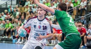 Ein Handballspieler im weißen Trikot hält den Ball, während er auf einem Hallenplatz von einem Spieler im grünen Trikot eng gedeckt wird; im Hintergrund beobachtet eine Zuschauermenge das Geschehen.