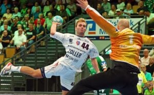 Ein Handballspieler im weißen Trikot springt zum Wurf hoch, während ein Torwart in Orange versucht, den Ball abzuwehren, und die Zuschauer von den Tribünen aus zuschauen.