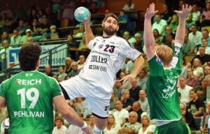 Ein Handballspieler in weißer Spielkleidung springt hoch, um einen Wurf anzusetzen, während zwei Verteidiger in grüner Spielkleidung versuchen, ihn zu blocken. Das Publikum verfolgt das Geschehen gebannt von den Tribünen aus.