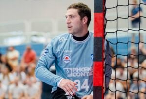 Ein männlicher Handballtorwart in einem hellblauen Trikot steht während eines Spiels in der Nähe des Torpfostens, im Hintergrund sind verschwommene Zuschauer zu sehen.