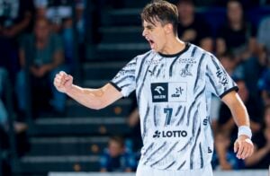 Ein männlicher Handballspieler in einem schwarz-weißen Trikot jubelt laut und reckt triumphierend die Faust in die Höhe auf einem Hallenfeld, während die Zuschauer im Hintergrund verschwommen zu sehen sind.