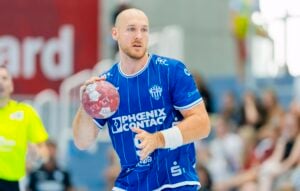 Ein männlicher Handballspieler in einem blauen Trikot hält einen Ball in der Hand und macht sich bereit zum Wurf. Im Hintergrund sind verschwommene Zuschauer und ein Schiedsrichter in gelber Kleidung zu sehen, was auf ein Live-Spiel hindeutet.
