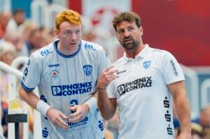 Zwei Männer in weißen Phoenix Contact-Sporttrikots stehen beieinander; der eine hört aufmerksam zu, während der andere gestikuliert und möglicherweise Anweisungen gibt – das Ganze spielt sich bei einer Hallensportveranstaltung ab.