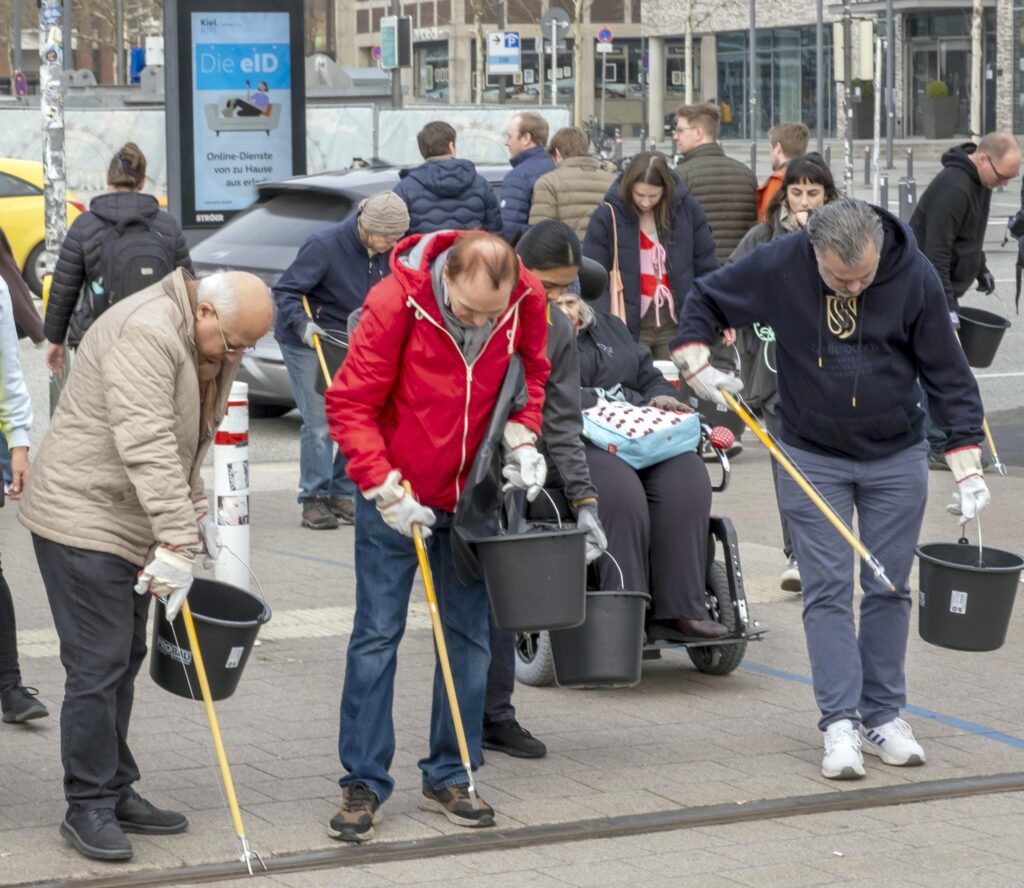 Eine Gruppe von Menschen in Jacken und Handschuhen säubert im Rahmen des THW-Kiel-Projekts „För De Küste – Nachhaltigkeit“ mit Müllgreifern und Eimern einen städtischen Gehweg. Im Hintergrund sind andere zu sehen, die spazieren gehen oder zuschauen, während im Hintergrund Gebäude und Fahrzeuge zu erkennen sind.