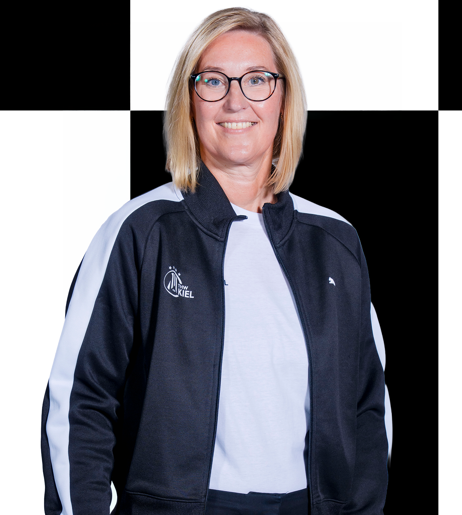 Eine Frau mit blonden Haaren und Brille, die eine schwarz-weiße Sportjacke über einem weißen Hemd trägt, lächelt vor einem schwarz-weiß karierten Hintergrund – ein perfektes Abbild der Mitarbeiter der THW Kiel-Geschäftsstelle. Für Kontaktdaten oder weitere Informationen wenden Sie sich bitte direkt an unser Büroteam.