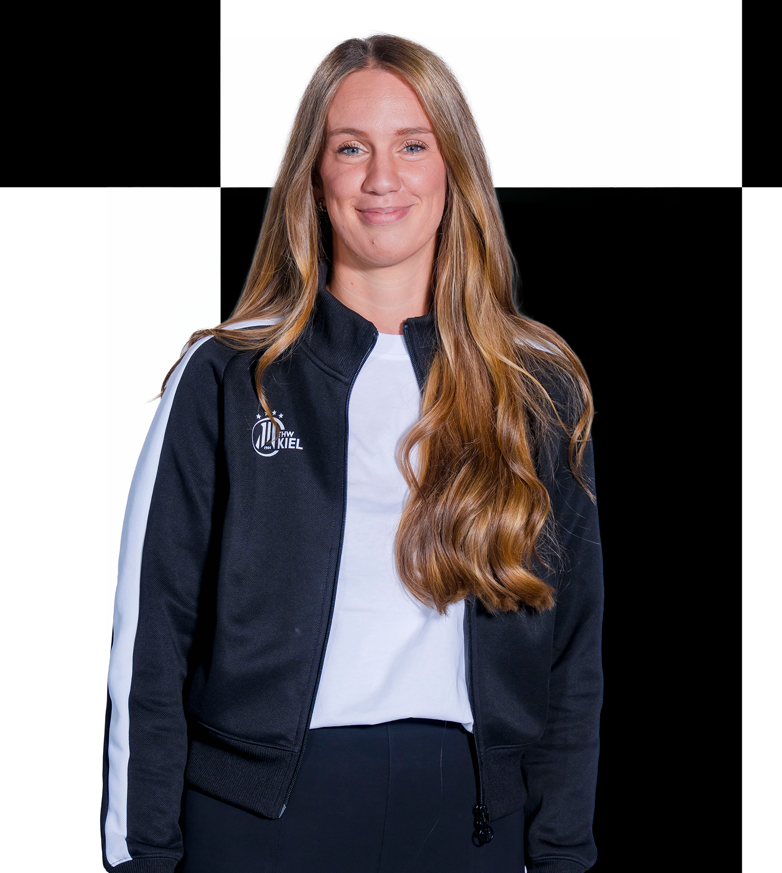 Eine Frau mit langen, welligen blonden Haaren, die eine schwarz-weiße Sportjacke trägt, steht lächelnd vor einem schwarz-weiß karierten Hintergrund und verkörpert damit den professionellen Geist der Mitarbeiter der THW Kiel-Geschäftsstelle. Für weitere Informationen oder zur Kontaktaufnahme nutzen Sie bitte die angegebenen Kontaktdaten.