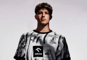 Ein junger Mensch in einem schwarz-weißen Sporttrikot mit verschiedenen Logos steht vor einem hellen, neutralen Hintergrund und blickt selbstbewusst in die Kamera.