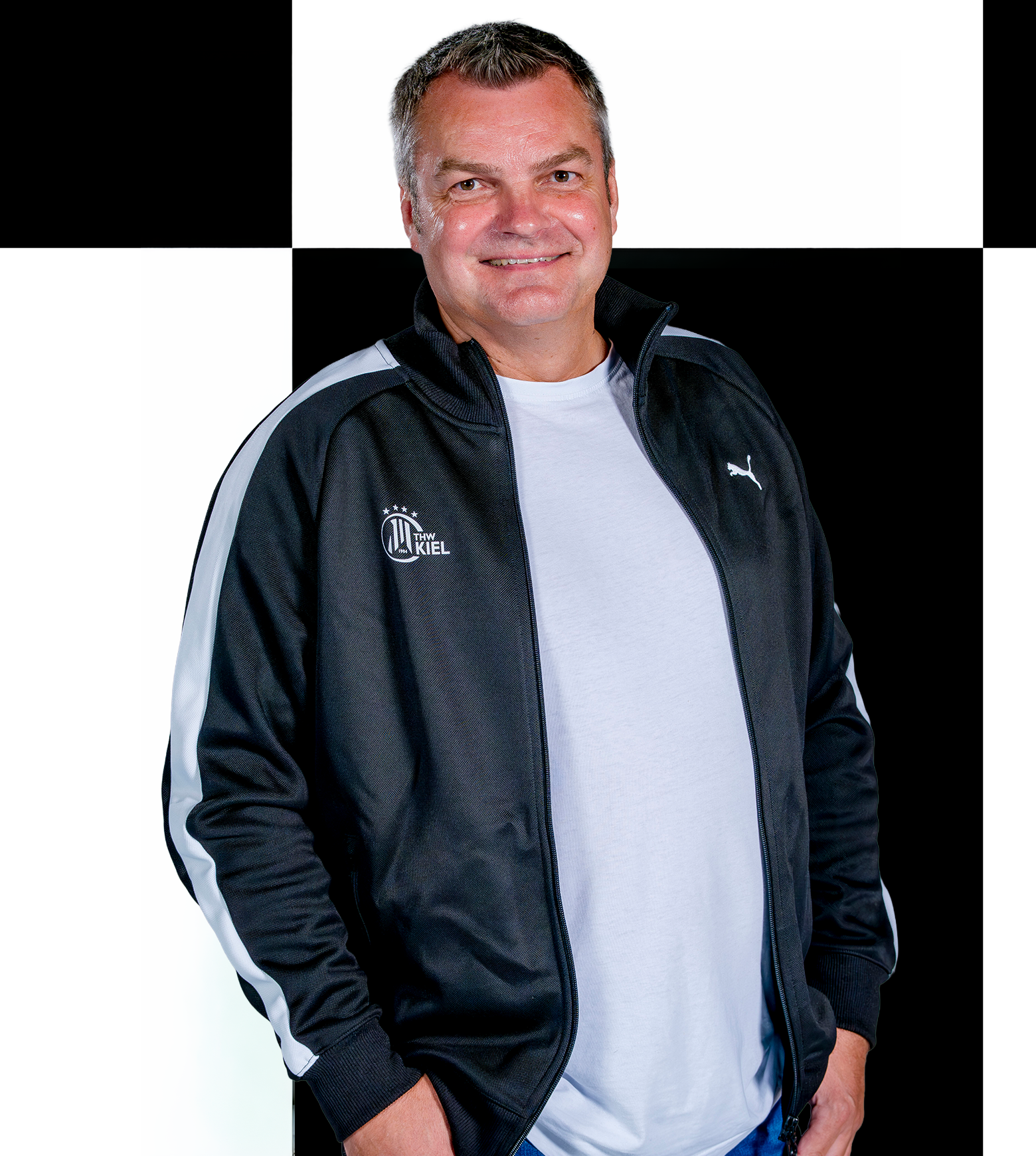 Björn Ein Mann in einer schwarzen Puma-Jacke mit weißen Streifen und einem weißen T-Shirt steht lächelnd vor einem schwarz-weiß karierten Hintergrund und repräsentiert den THW Kiel als Ansprechpartner für Sponsoren und Partner.