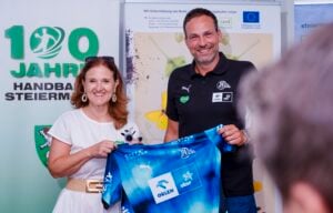 Eine Frau und ein Mann stehen lächelnd nebeneinander und halten ein blaues Sporttrikot in den Händen. Sie befinden sich bei einer Veranstaltung, hinter ihnen sind Transparente zu sehen, darunter eines mit der Aufschrift „100 Jahre Handball Steiermark“.