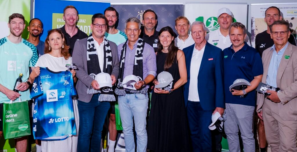 HANDBALL 2026/27: THW Kiel im Trainingslager in Graz Eine Gruppe von Menschen, von denen einige Schutzhelme und Trikots in den Händen halten, steht beieinander und lächelt für ein Foto bei einer Hallenveranstaltung, im Hintergrund sind Sport- und Sponsorenbanner zu sehen.