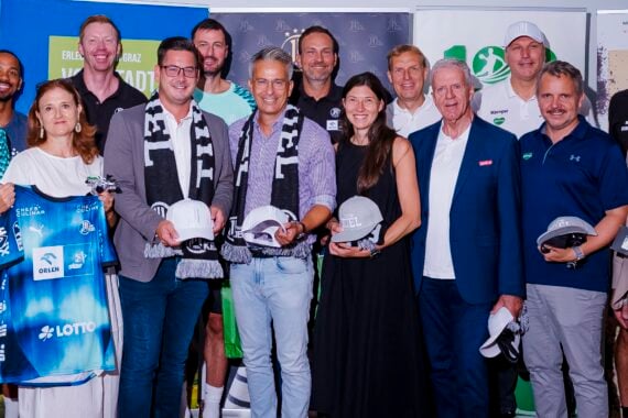 Eine Gruppe von Menschen, von denen einige Schutzhelme und Trikots in den Händen halten, steht beieinander und lächelt für ein Foto bei einer Hallenveranstaltung, im Hintergrund sind Sport- und Sponsorenbanner zu sehen.