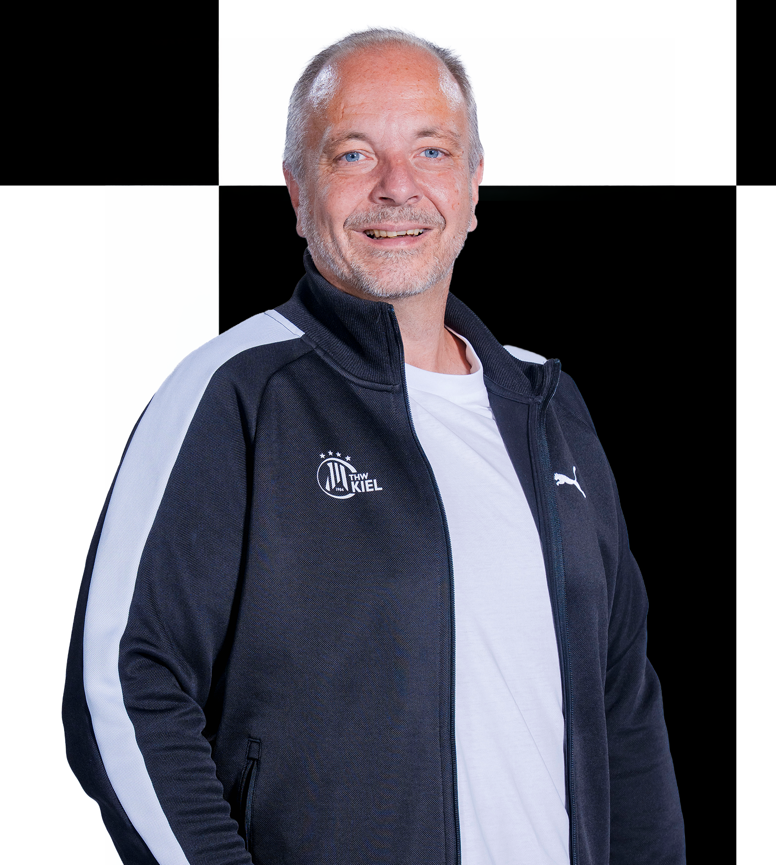 Ein lächelnder Mann mit kurzen grauen Haaren, der offenbar ein Mitarbeiter der THW Kiel-Geschäftsstelle ist, trägt ein schwarz-weißes Sakko über einem weißen Hemd und steht vor einem schwarz-weiß karierten Hintergrund. Die Kontaktdaten finden Sie im Teamverzeichnis.