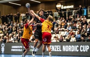 Ein Handballspieler in schwarzer Kleidung springt zum Wurf hoch, während zwei Spieler in gelber und roter Kleidung verteidigen. Im Hintergrund ist eine mit Zuschauern gefüllte Halle sowie ein „Food & Beverage“-Schild zu sehen.