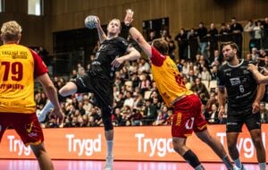 Ein Handballspieler in schwarzer Kleidung springt zum Wurf ab, während ein Verteidiger in gelber Kleidung versucht, den Wurf zu blocken. Andere Spieler und eine Menge Zuschauer verfolgen das Geschehen gespannt im Hintergrund.