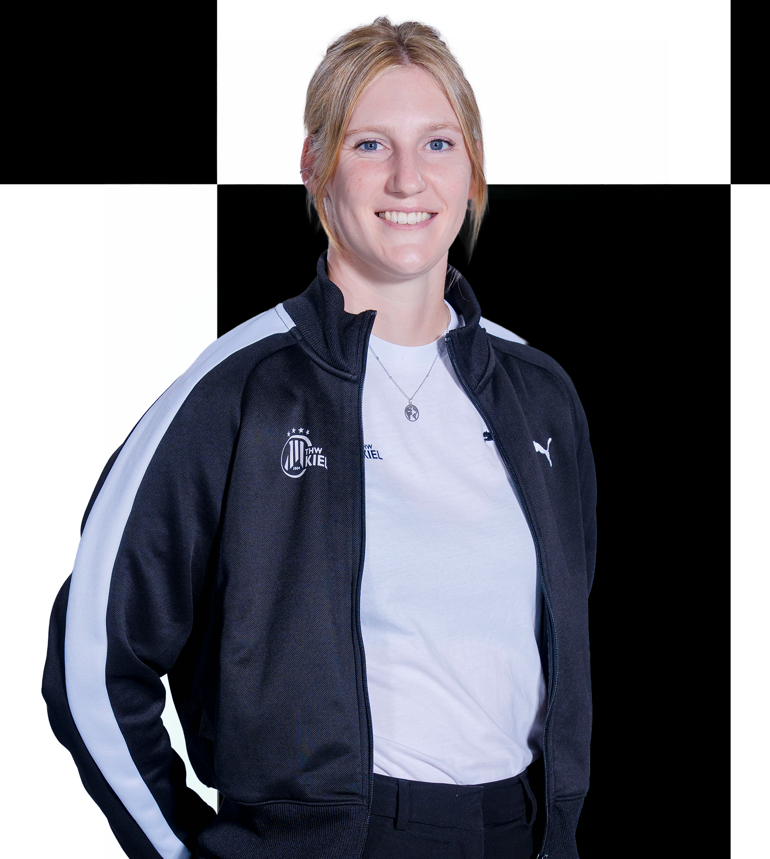 Eine Frau mit blonden Haaren, die eine schwarz-weiße Puma-Jacke über einem weißen Hemd trägt, steht lächelnd vor einem schwarz-weiß karierten Hintergrund und verkörpert die freundliche Atmosphäre von THW Kiel – Geschäftsstelle, Mitarbeiter, Kontakt.
