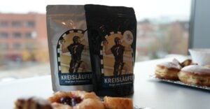 Zwei Tüten „Kreisläufer“-Kaffee mit der Abbildung eines Handballspielers auf einem Tisch, im Hintergrund sind Gebäck und verschwommene Stadtgebäude zu sehen.