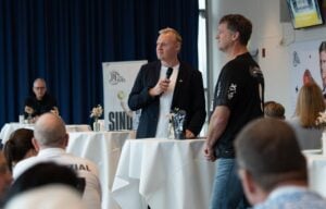 Ein Mann im Anzug spricht an einem Stehtisch in ein Mikrofon, während ein anderer Mann im T-Shirt ihm zuhört. Die Menschen sitzen in einem modernen, gut beleuchteten Raum mit blauen Vorhängen und Postern an den Wänden und schauen zu.