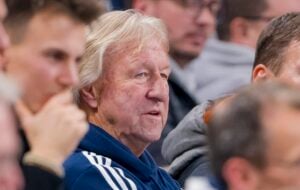 Ein älterer Mann mit hellem Haar, der eine blau-weiße Jacke trägt, sitzt inmitten einer Gruppe von Menschen und scheint sich auf etwas zu konzentrieren, das sich außerhalb des Bildausschnitts befindet.
