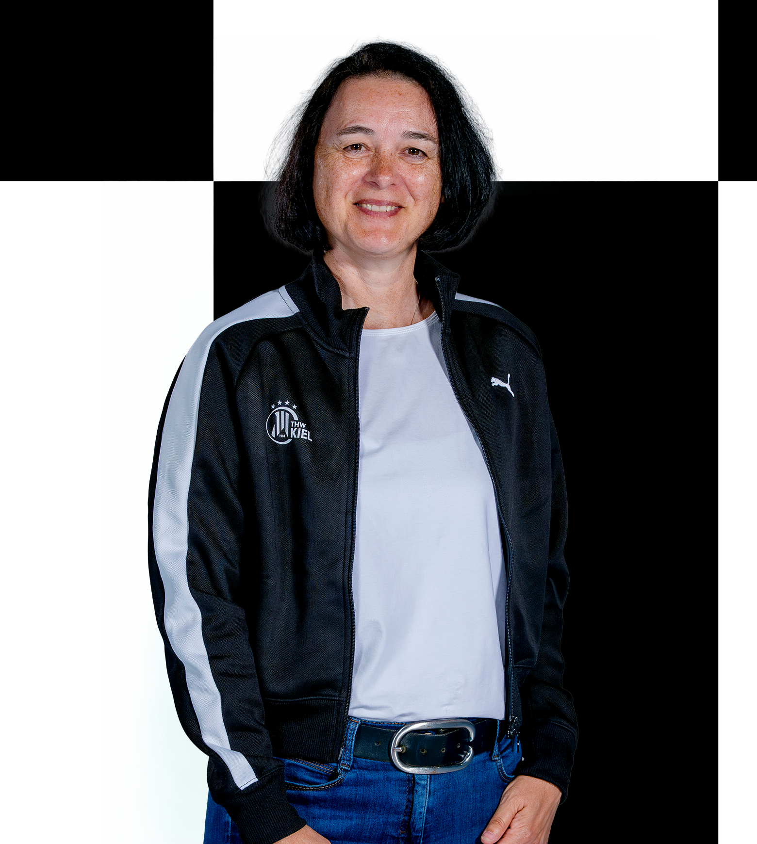 Eine Frau mit dunklem Haar, die eine schwarz-weiße Puma-Jacke und Jeans trägt, steht lächelnd vor einem schwarz-weiß karierten Hintergrund, der an die Geschäftsstelle des THW Kiel erinnert. Informationen zum Mitarbeiterkontakt entnehmen Sie bitte den offiziellen Quellen.