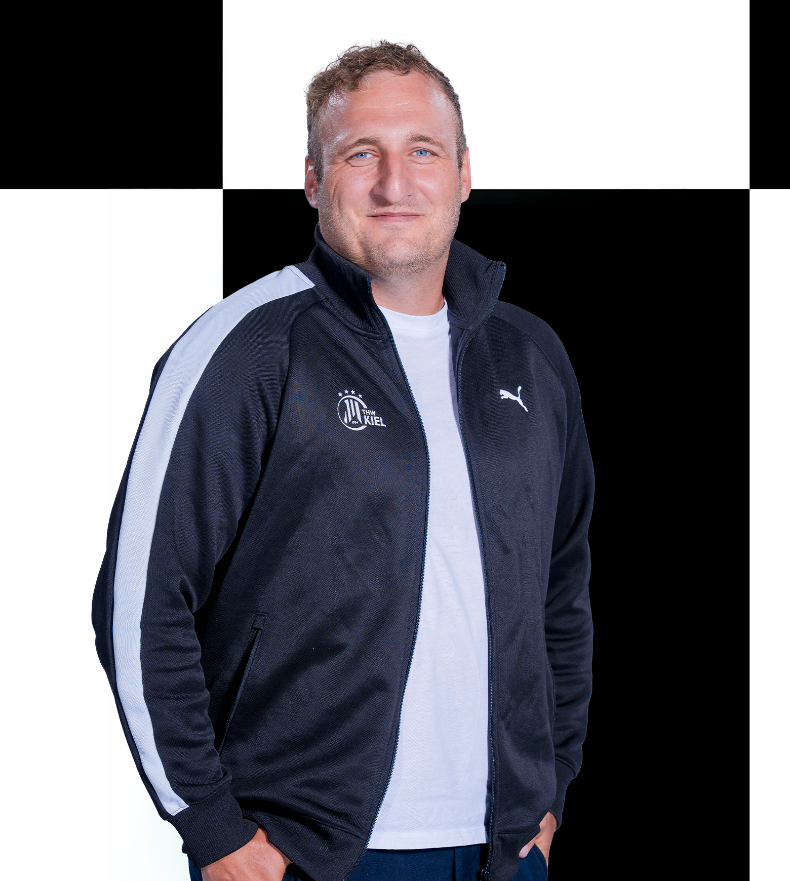 Marvin Ein Mann in einer schwarz-weißen Puma-Jacke steht vor einem schwarz-weiß karierten Hintergrund, lächelt und hat die Hände in den Taschen – er verkörpert perfekt den Stil des THW Kiel, für den Sportfive stolz als Ansprechpartner für Sponsoren und Partner fungiert.