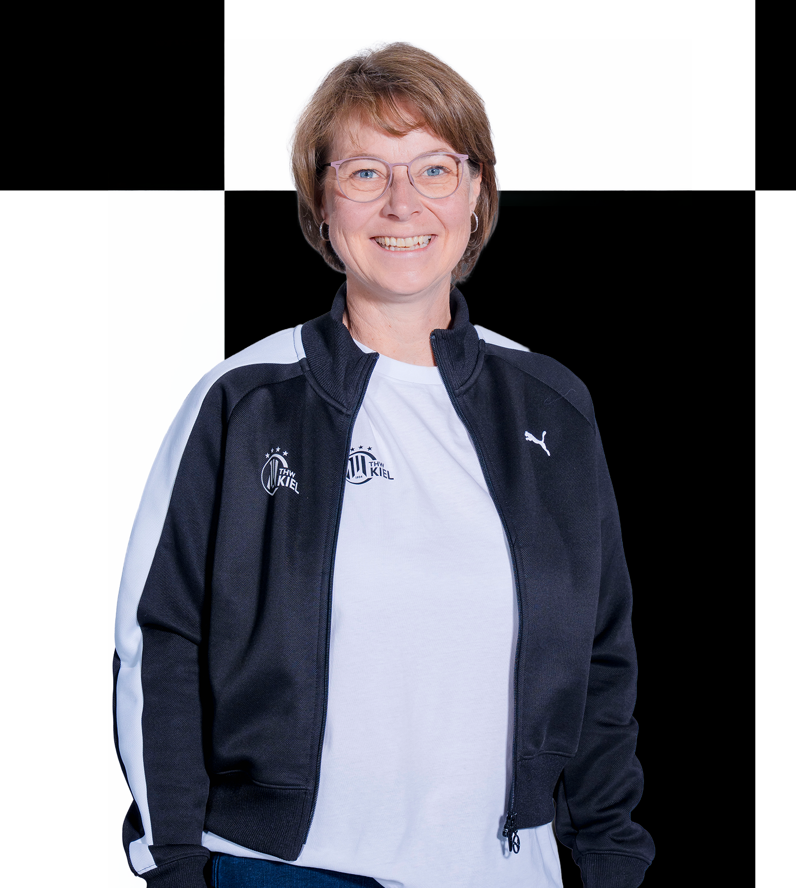 Eine lächelnde Frau mit kurzen Haaren und Brille, die bei der THW Kiel-Geschäftsstelle arbeitet, trägt eine schwarz-weiße Puma-Jacke über einem weißen Hemd und steht vor einem schwarz-weiß karierten Hintergrund. Die Kontaktdaten finden Sie auf der offiziellen Website.