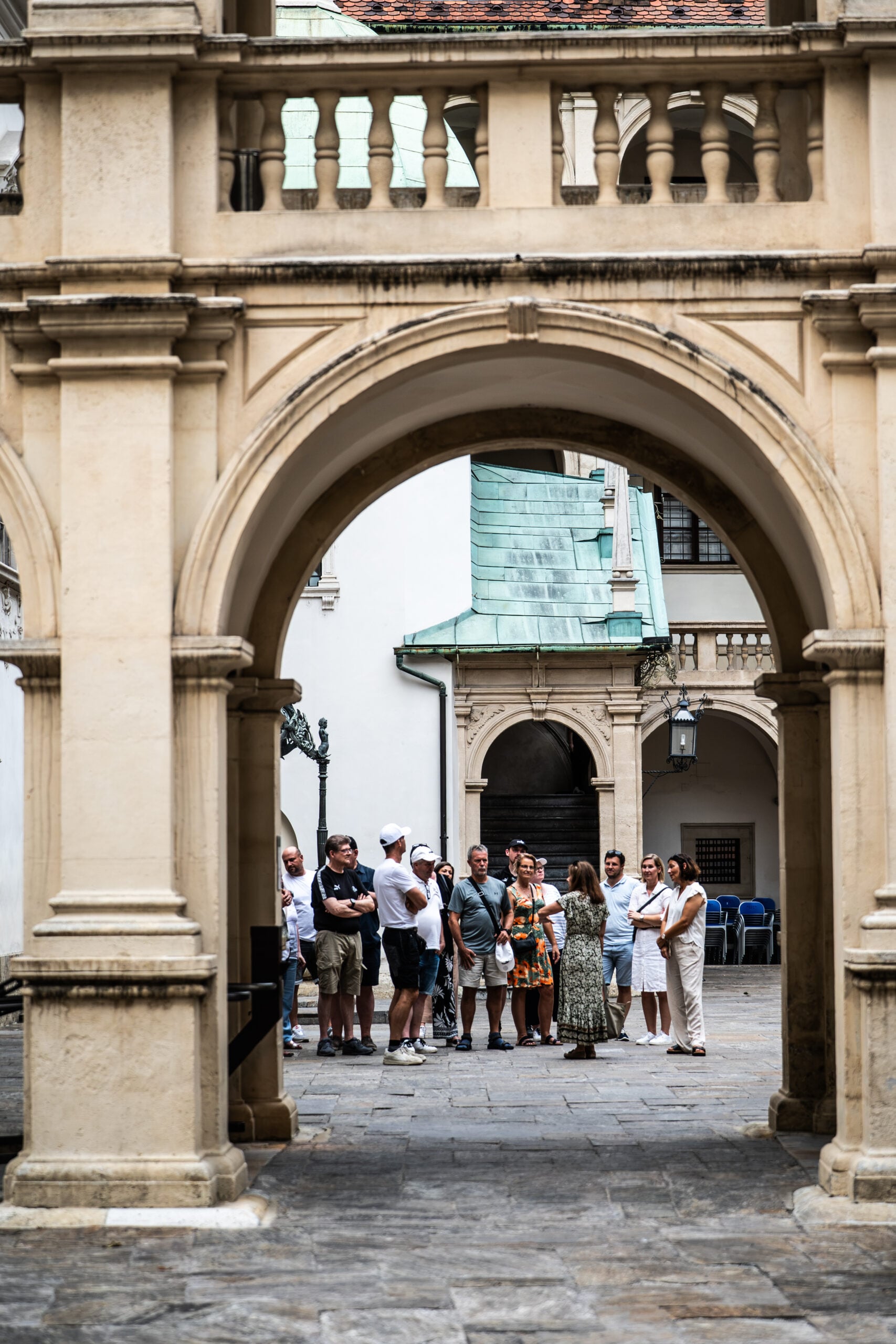 Eine Gruppe von Touristen steht gemeinsam unter einem steinernen Torbogen in einem historischen Innenhof und lauscht einem Reiseleiter. Das Gebäude hat helle Wände, gewölbte Öffnungen und ein grün patiniertes Dach – ganz ähnlich wie die Kulisse, vor der Neu-Trainer Börge Lund beim THW Kiel vorgestellt wurde.