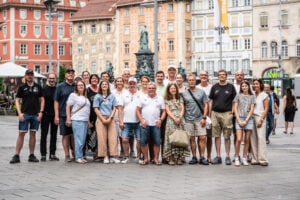 Eine große Gruppe von Erwachsenen posiert gemeinsam im Freien auf einem Stadtplatz im europäischen Stil und lächelt in die Kamera. Im Hintergrund sind Gebäude mit bunten Fassaden und eine Statue zu sehen, die an Szenen erinnern, in denen der neue Trainer Börge Lund beim THW Kiel vorgestellt wurde.