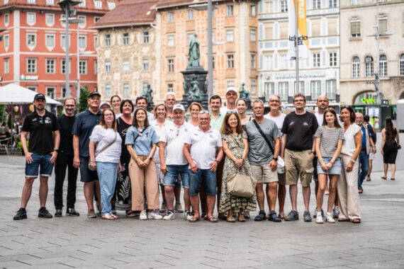 Eine große Gruppe von Erwachsenen posiert gemeinsam im Freien auf einem Stadtplatz im europäischen Stil und lächelt in die Kamera. Im Hintergrund sind Gebäude mit bunten Fassaden und eine Statue zu sehen, die an Szenen erinnern, in denen der neue Trainer Börge Lund beim THW Kiel vorgestellt wurde.