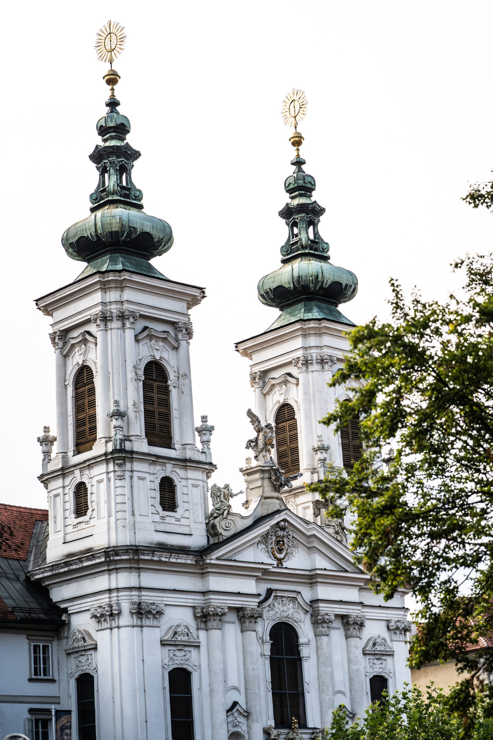 Eine Kirche im Barockstil mit zwei kunstvoll verzierten Türmen, grünen Kuppeln und goldenen Kreuzen. Die Fassade besticht durch Bogenfenster und dekorative Details. Sie erinnert an die Pracht, die zu sehen war, als Neu-Trainer Börge Lund beim THW Kiel vorgestellt wurde; auf der rechten Seite ist teilweise ein Baum zu erkennen.