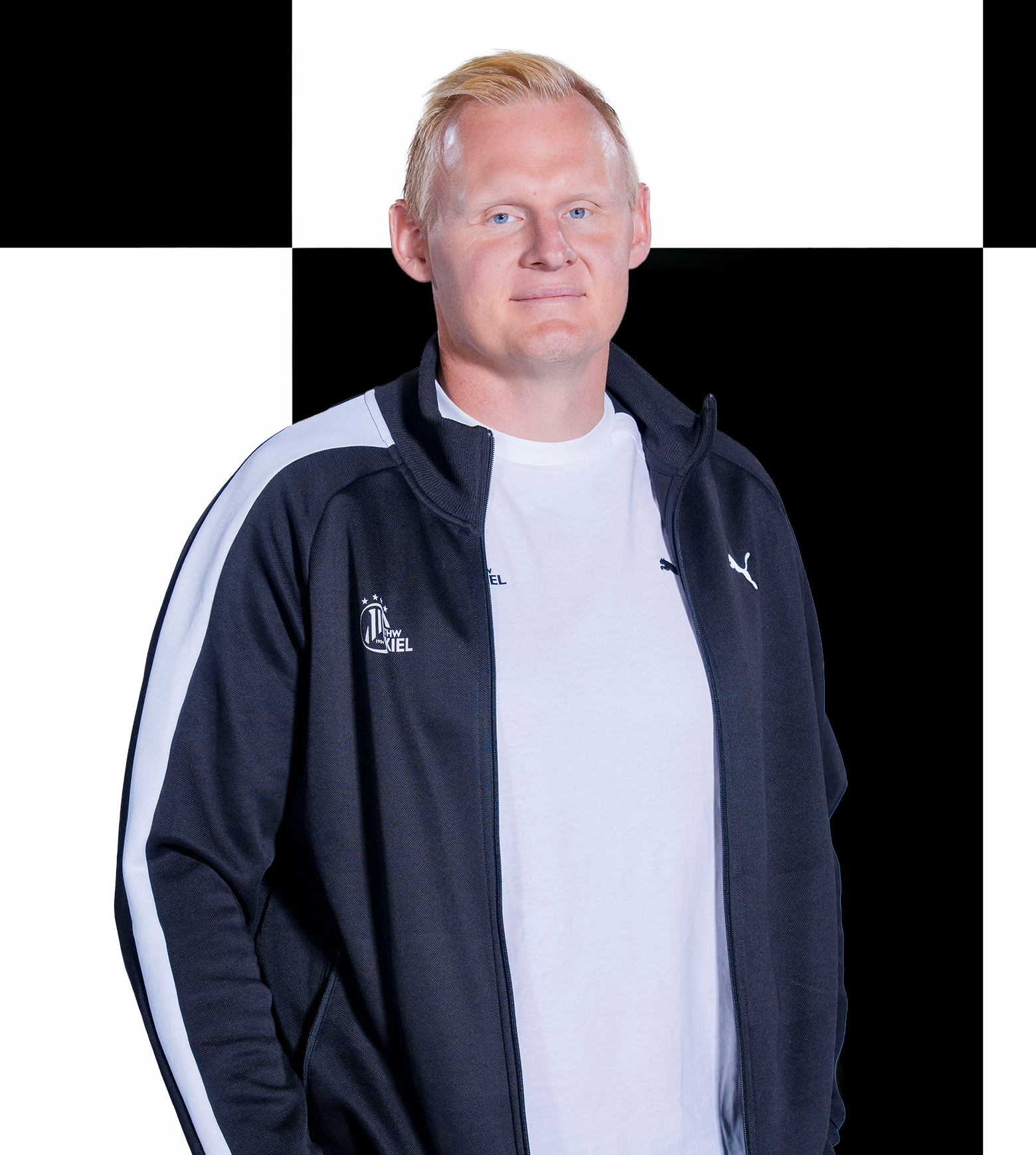 Eine Person mit kurzen blonden Haaren, die ein weißes Hemd und eine schwarz-weiße Jacke trägt, steht vor einem schwarz-weiß karierten Hintergrund, der an die Geschäftsstelle des THW Kiel erinnert. Dieses Bild könnte ohne Weiteres einen ihrer Mitarbeiter darstellen, der bereit ist, bei allen Kontaktanfragen zu helfen.