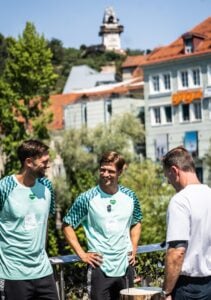 Drei Männer – zwei in identischen blaugrünen Sporthemden und einer in einem weißen Hemd – stehen draußen und unterhalten sich. Im sonnigen Hintergrund sind bunte Gebäude und ein Uhrturm zu sehen.