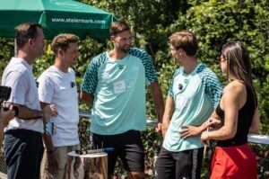 An einem sonnigen Tag stehen fünf Personen im Freien und unterhalten sich. Unter ihnen befinden sich drei Stars aus Graz sowie zwei Handballstars aus Kiel beim Steiermark-Test – zwei Männer in einheitlichen grünen Sporttrikots, während die anderen Freizeitkleidung tragen. Sie haben sich um einen Holztisch versammelt, auf dem ein Hammer und große Nägel liegen, im Hintergrund sind Bäume und ein grüner Sonnenschirm zu sehen.