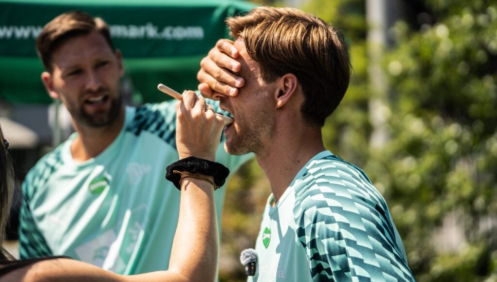 Rune_Harry-6_Titel Eine Person trägt mit einem Pinsel etwas auf das Gesicht eines Mannes auf, während dieser sich die Augen zuhält; daneben steht im Freien ein weiterer Mann in passender Sportkleidung – vielleicht Teilnehmer der Veranstaltung „Drei Stars aus Graz plus zwei Kieler Handball-Stars beim Steiermark-Test“, die sich auf die Veranstaltung vorbereiten.