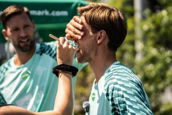 Eine Person trägt mit einem Pinsel etwas auf das Gesicht eines Mannes auf, während dieser sich die Augen zuhält; daneben steht im Freien ein weiterer Mann in passender Sportkleidung – vielleicht Teilnehmer der Veranstaltung „Drei Stars aus Graz plus zwei Kieler Handball-Stars beim Steiermark-Test“, die sich auf die Veranstaltung vorbereiten.