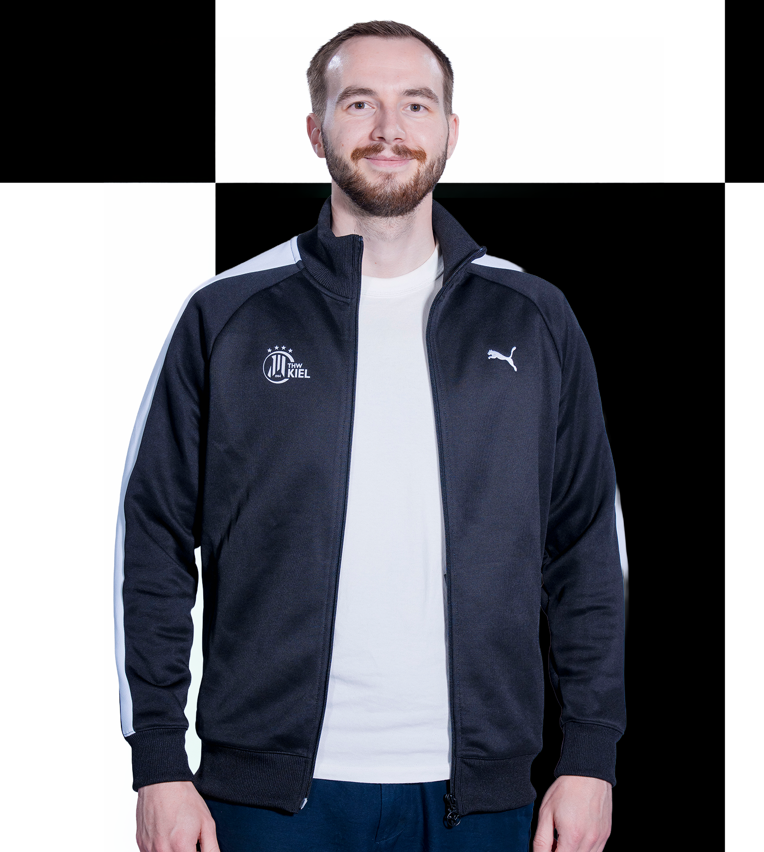 Silva Ein Mann mit kurzen Haaren und Bart lächelt. Er trägt eine schwarze Trainingsjacke mit weißen Streifen und THW-Kiel-Logos und steht vor einem schwarz-weiß karierten Hintergrund – was seine Rolle als Ansprechpartner für Sponsoren und Sportfive-Partner verdeutlicht.