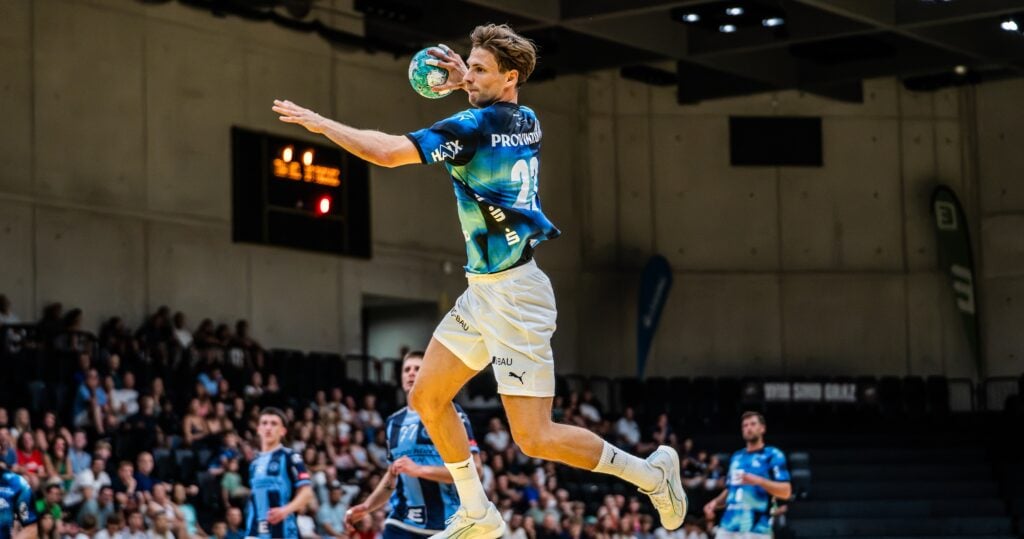 Spiel-12 Ein männlicher Handballspieler springt in die Luft und macht sich bereit, den Ball während eines Spiels in einer Halle zu werfen, während im Hintergrund Zuschauer zuschauen.