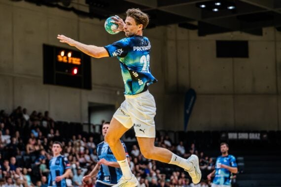 Ein männlicher Handballspieler springt in die Luft und macht sich bereit, den Ball während eines Spiels in einer Halle zu werfen, während im Hintergrund Zuschauer zuschauen.