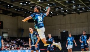 Ein männlicher Handballspieler in einem blauen Trikot springt in die Luft, um den Ball ins Tor zu werfen, während die Verteidiger zusehen. Im Hintergrund sitzen Zuschauer unter heller Hallenbeleuchtung.