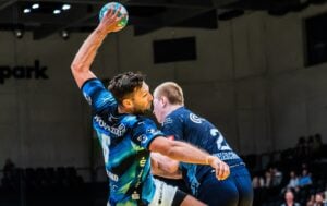 Ein Handballspieler in einem blauen Trikot hebt den Ball zum Wurf an, während er von einem anderen Spieler eng gedeckt wird; beide sind auf einem Hallenfeld im Einsatz.