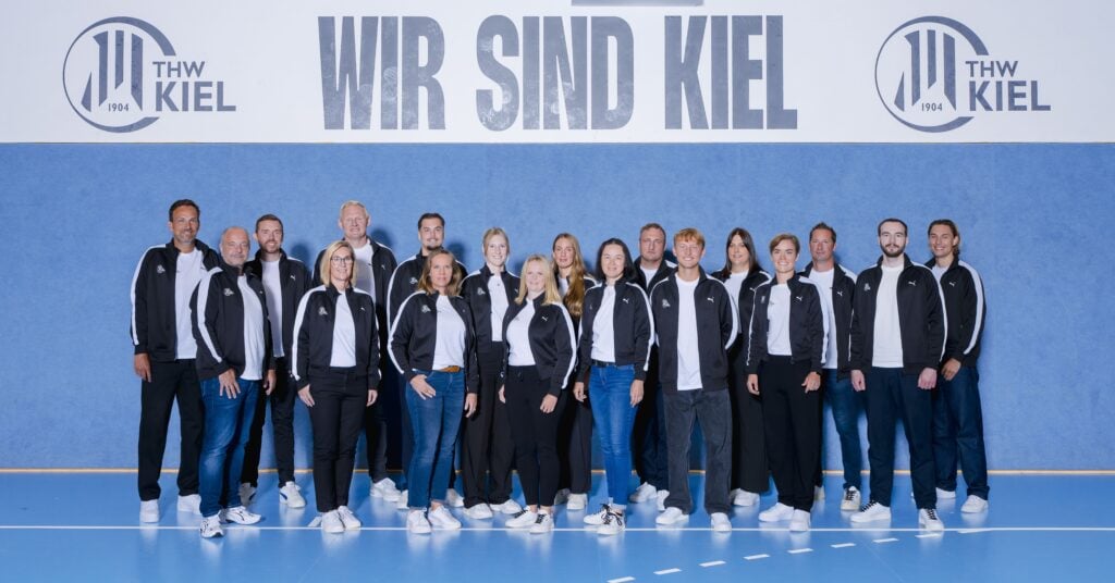 Vor einer blauen Wand, auf der „WIR SIND KIEL“ und das THW-Kiel-Logo aufgedruckt sind, steht eine Gruppe von Menschen in einheitlichen schwarz-weißen Jacken – sie stehen für die engagierten Mitarbeiter der THW-Kiel-Geschäftsstelle, die jederzeit für Kontaktaufnahmen zur Verfügung stehen.