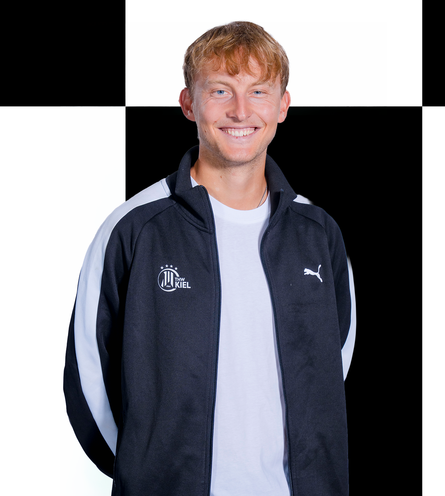 Eine lächelnde Person, die eine schwarz-weiße Puma-Sportjacke über einem weißen Hemd trägt, steht vor einem schwarz-weiß karierten Hintergrund und verkörpert damit den professionellen Geist, der bei den Mitarbeitern der THW Kiel-Geschäftsstelle oft zu beobachten ist.