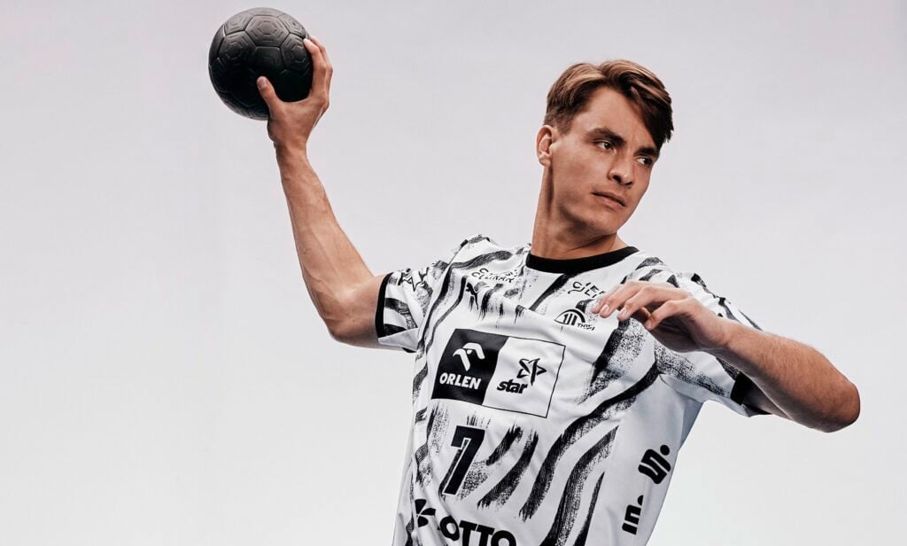 Titel_Koester_Wurf Ein männlicher Handballspieler in einem schwarz-weißen Trikot macht sich bereit, einen schwarzen Handball zu werfen. Vor einem schlichten Hintergrund wirkt er konzentriert, während er den Arm hinter dem Kopf erhoben hält.