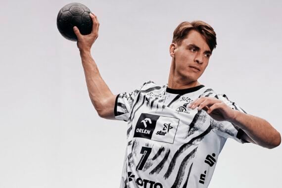 Ein männlicher Handballspieler in einem schwarz-weißen Trikot macht sich bereit, einen schwarzen Handball zu werfen. Vor einem schlichten Hintergrund wirkt er konzentriert, während er den Arm hinter dem Kopf erhoben hält.
